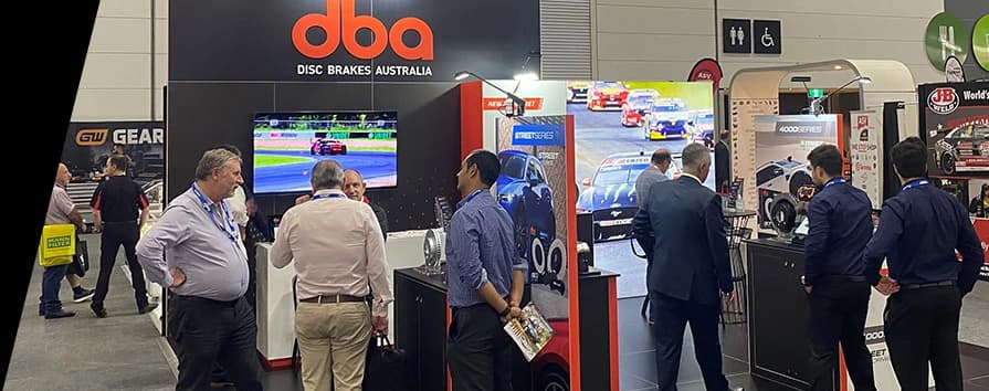 Stand espositivo DBA a una fiera dell'automobile con il logo aziendale in evidenza e partecipanti che osservano l'esposizione di prodotti e contenuti motorsport