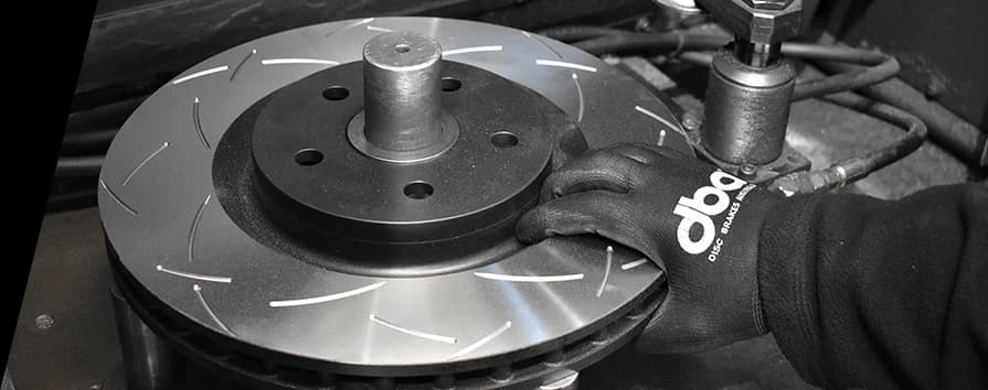 Installazione professionale di un disco freno scanalato ad alte prestazioni DBA da parte di un tecnico certificato con guanti a marchio Primo piano di un tecnico con guanti DBA che installa un disco freno scanalato ad alte prestazioni sul mozzo del veicolo durante il servizio freni