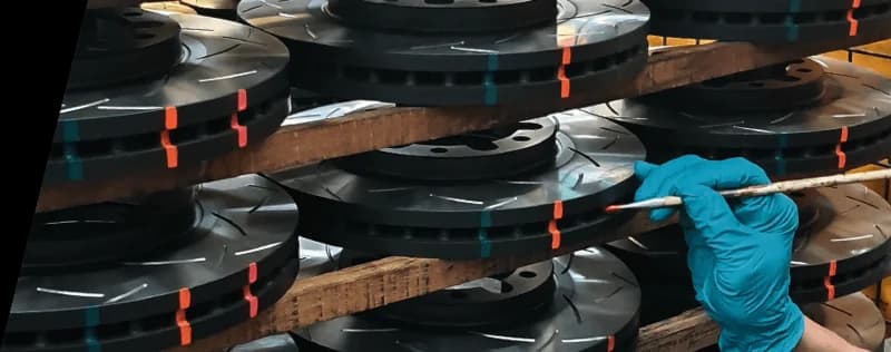 Ispezione del controllo qualità dei dischi freno DBA che mostra indicatori in vernice termica e motivi delle scanalature ingegnerizzati con precisione Tecnico con guanti blu che ispeziona dischi freno scanalati DBA impilati con contrassegni indicatori di temperatura colorati durante il processo di controllo qualità